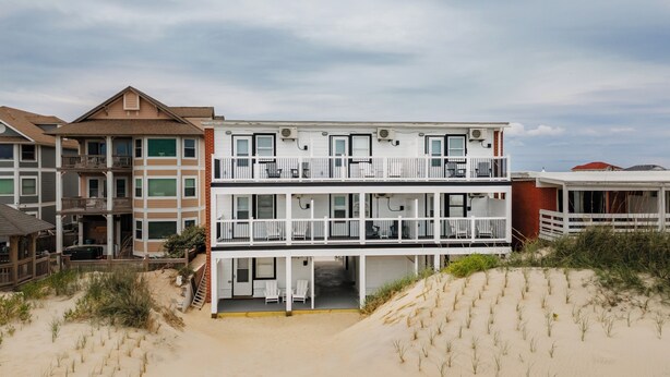 Un immeuble à plusieurs étages situé en bord de mer, doté de balcons, avec une plage de sable devant et un ciel dégagé.