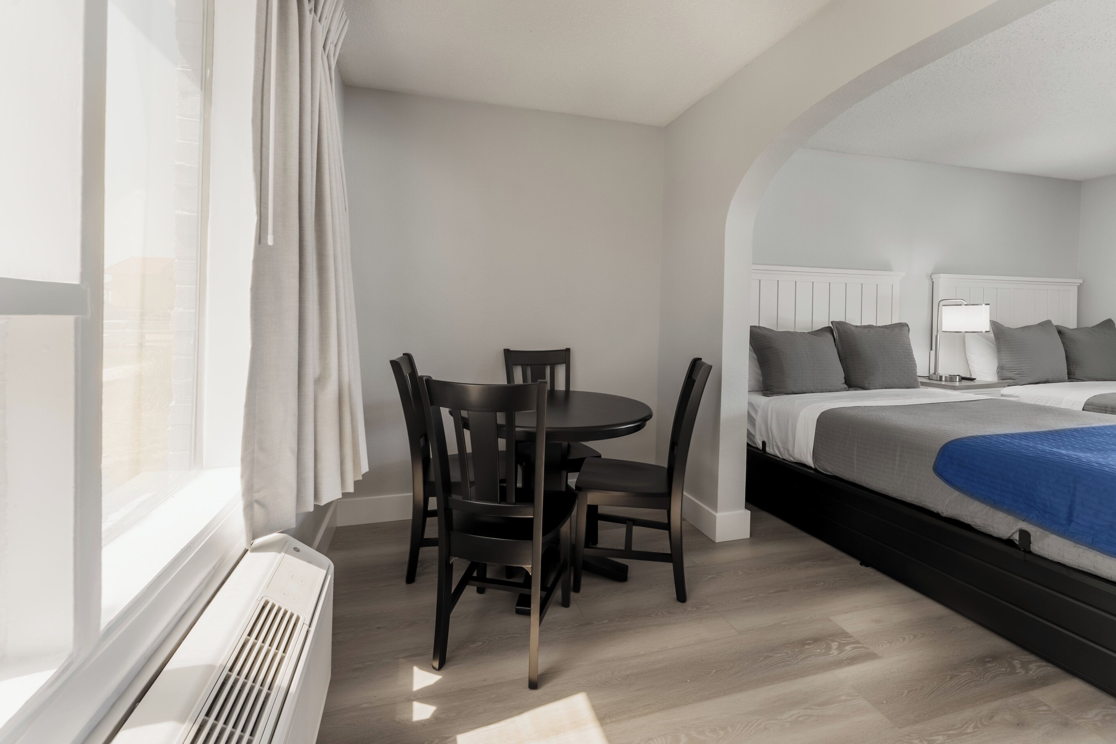 A modern hotel room with a bed, bedside table, lamp, and a dining area with chairs and a table.