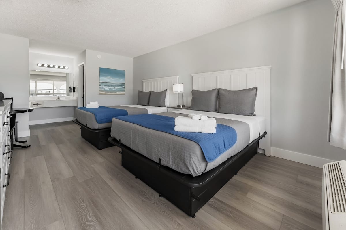A hotel room with two beds, a desk, a mirror, and a view of the sea.