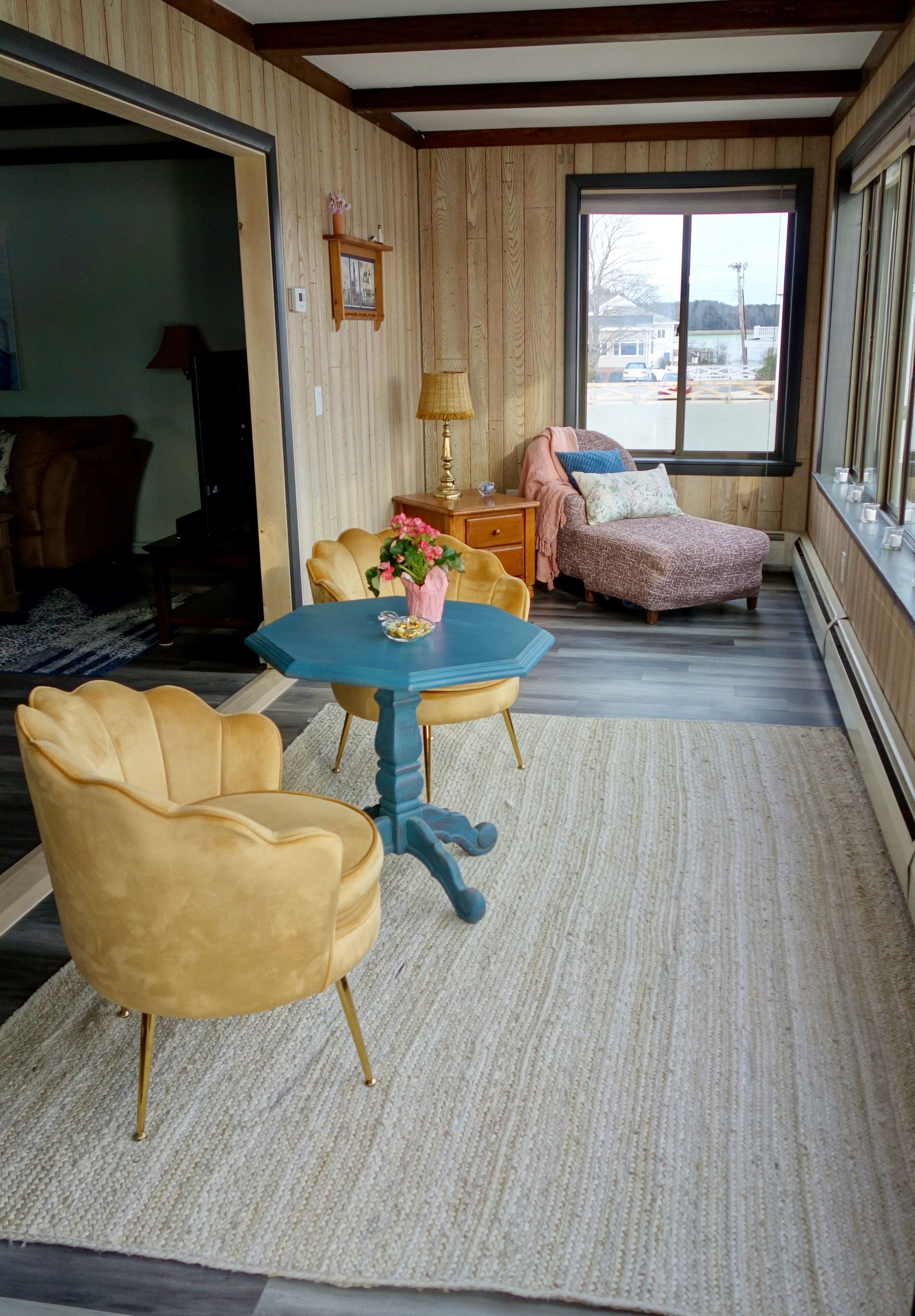 Una habitación acogedora con un revestimiento de madera, un ventanal grande, una mesa azul con sillas y un reposapiés rosa.