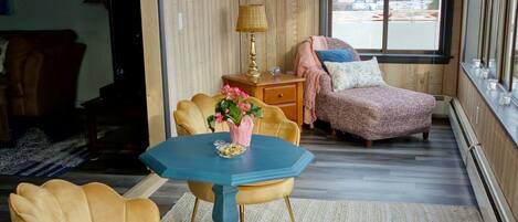 Una habitación acogedora con un revestimiento de madera, un ventanal grande, una mesa azul con sillas y un reposapiés rosa.