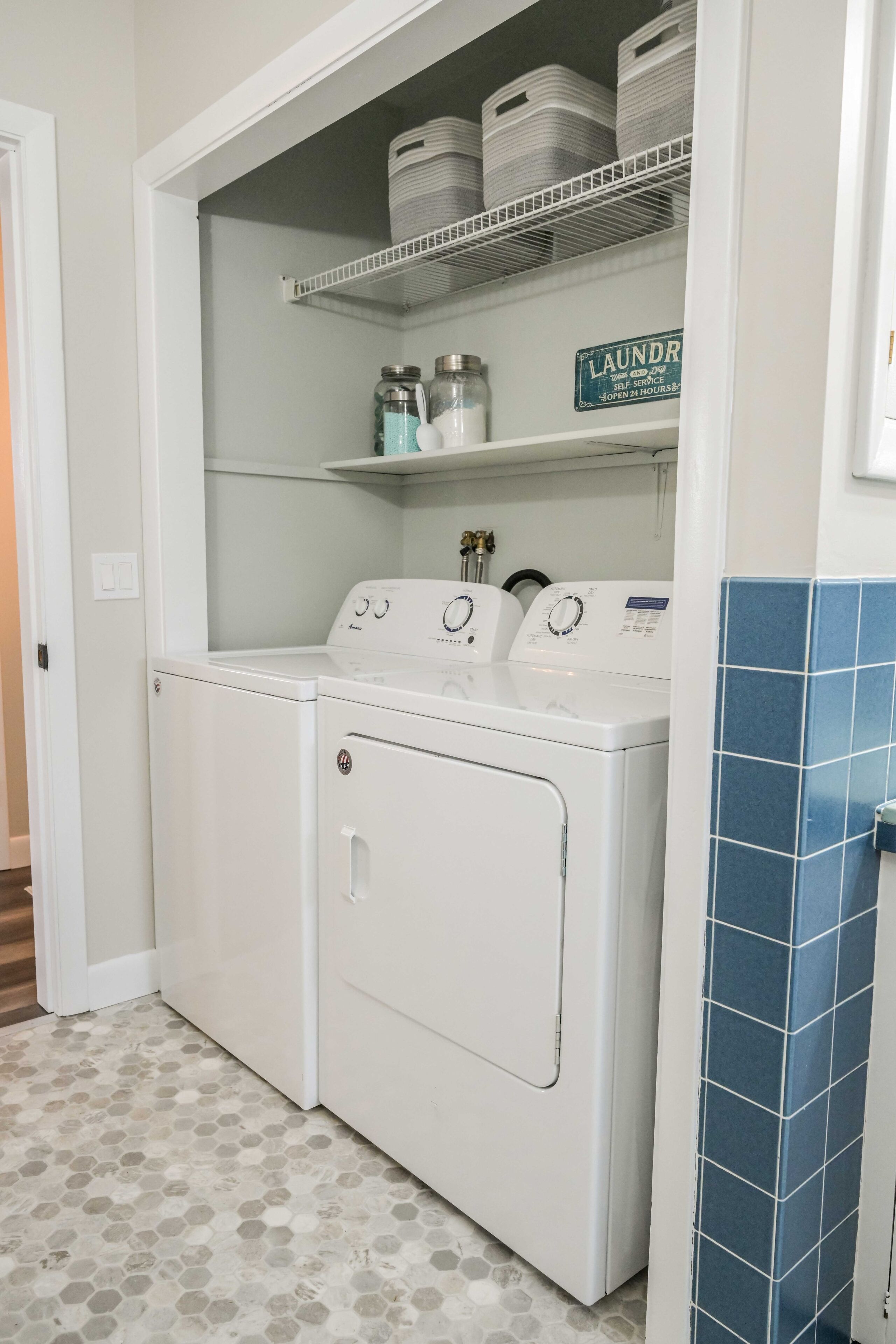 A laundry area with a front-loading washer and dryer, a shelf above with stacked laundry baskets, and a sign indicating self-service laundry hours.