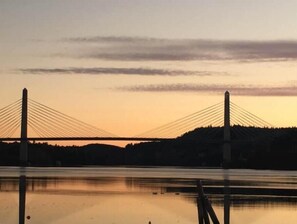 A suspension bridge at sunset with a calm water body in the foreground.