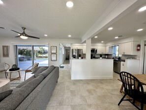Un vaste espace de vie comprenant un canapé gris, une table à manger avec des chaises, une cuisine avec des meubles blancs et un ventilateur de plafond.
