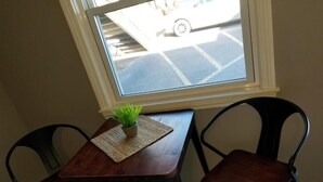 Un petit coin repas avec une table et deux chaises, une plante en pot posée sur la table et, par la fenêtre, une vue sur un parking.