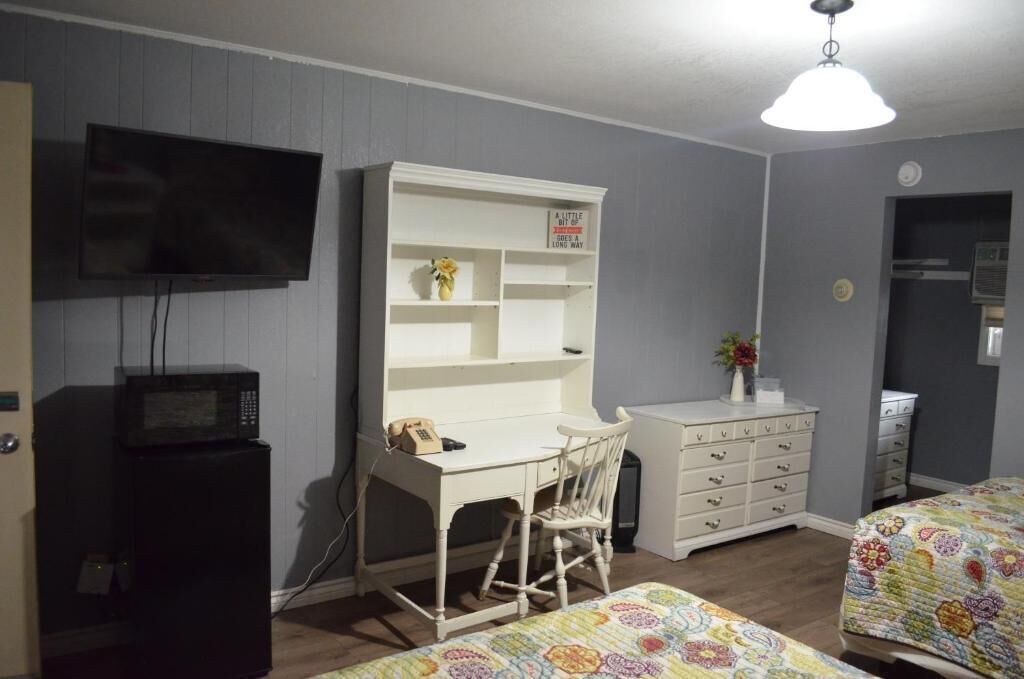 A room with a white desk, a television mounted on the wall, a microwave, a dresser, and two beds with colorful bedding.
