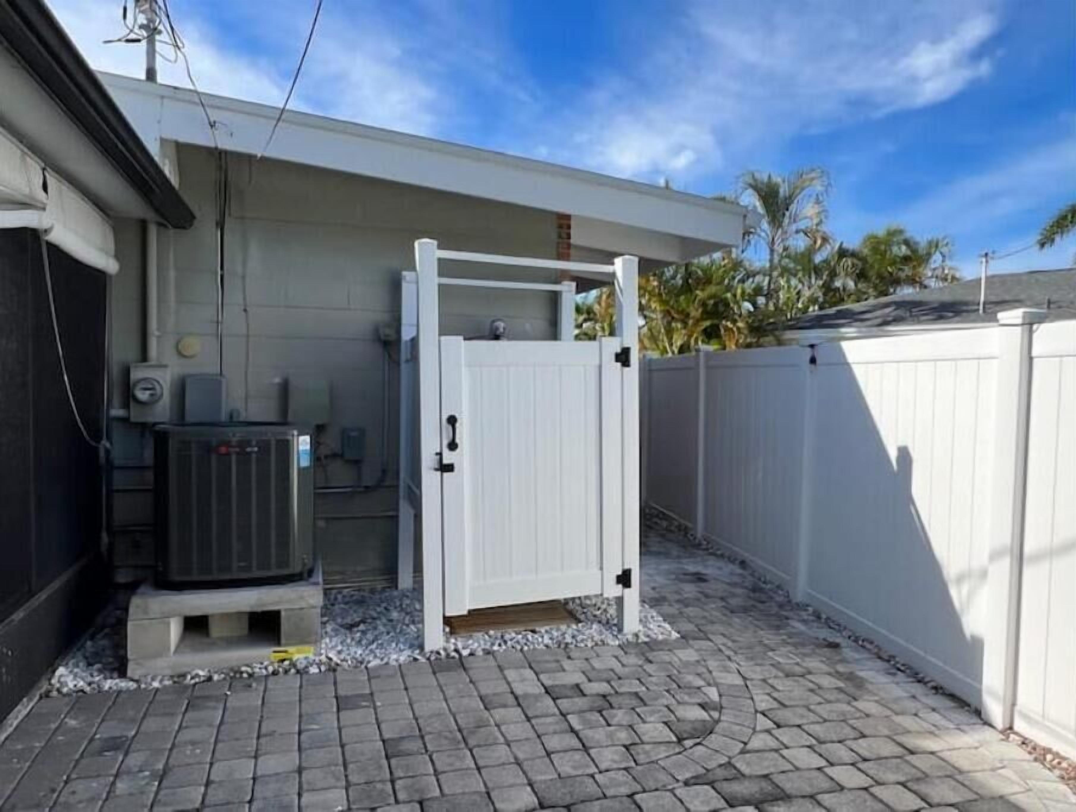 Un patio trasero cercado con un patio empedrado, una valla blanca y una zona de servicios con un equipo de aire acondicionado.
