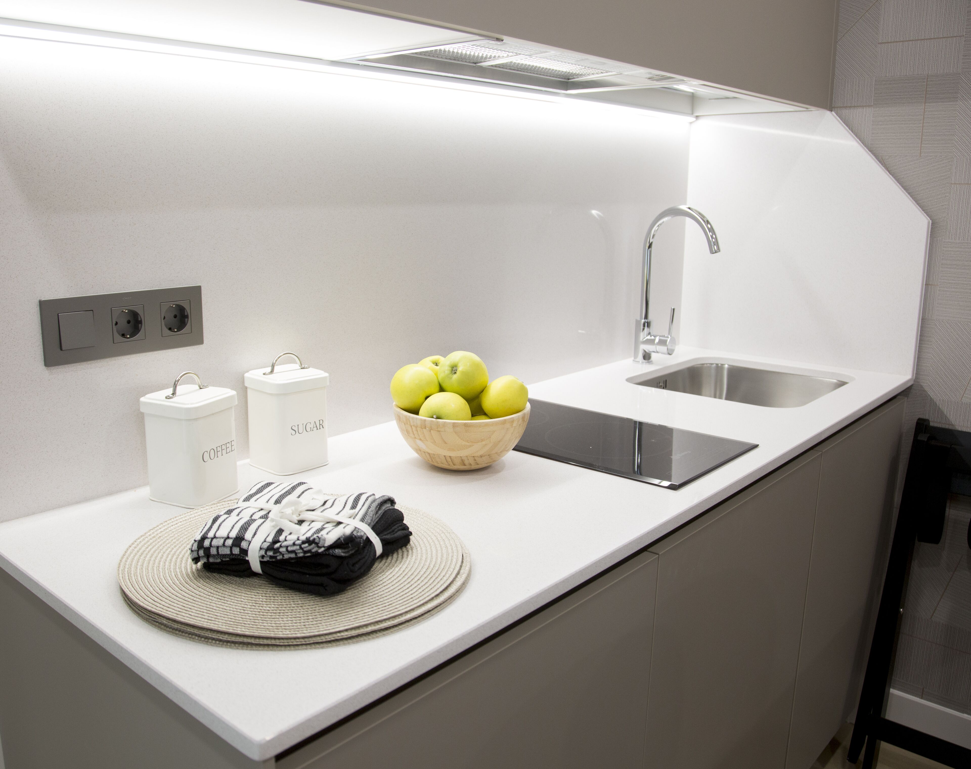 Cocina moderna con encimera blanca, un cuenco de manzanas, un recipiente para café y un azucarero.