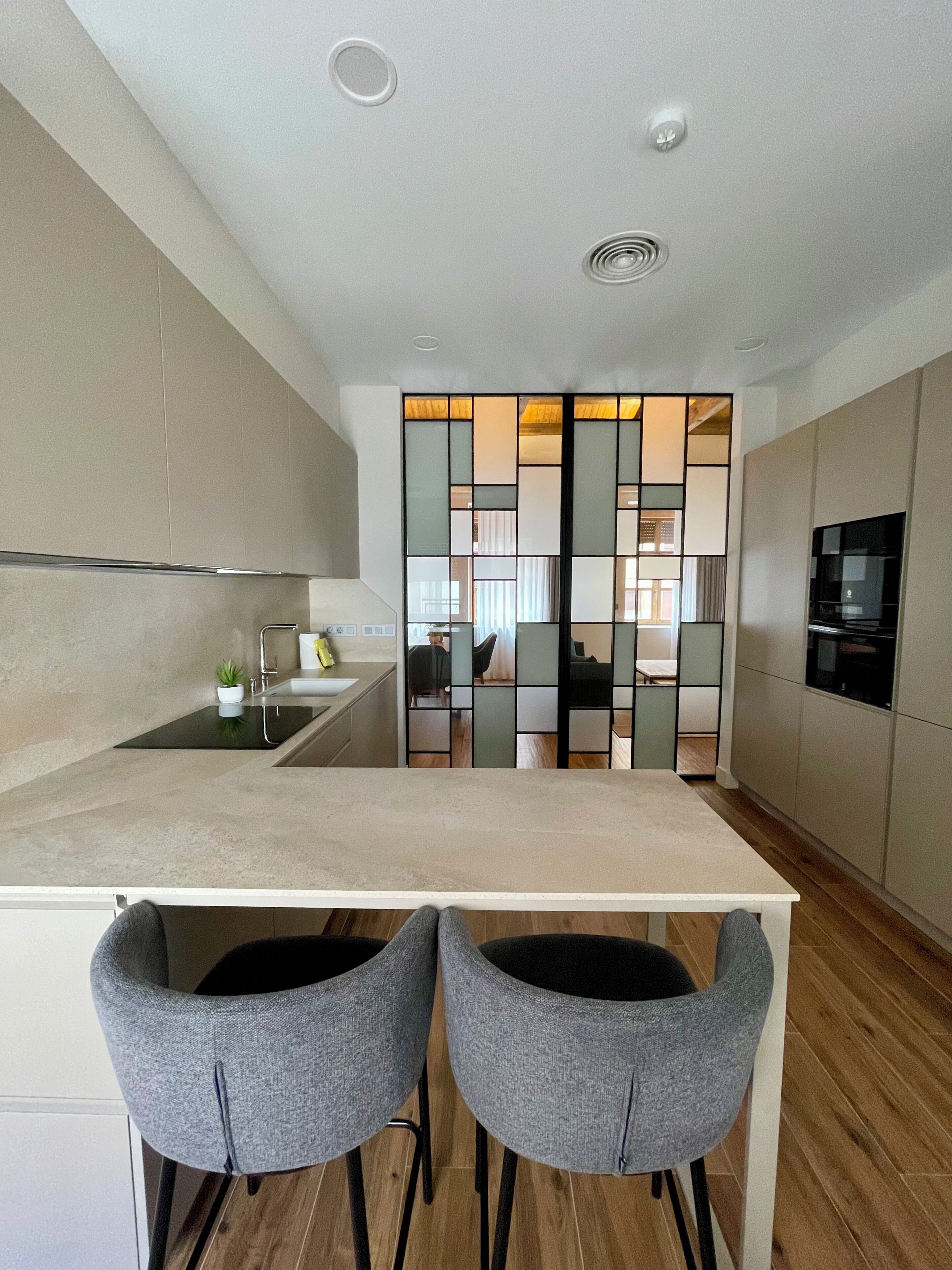 A modern kitchen with a central island, two grey chairs, and a view into the living area through a glass partition.
