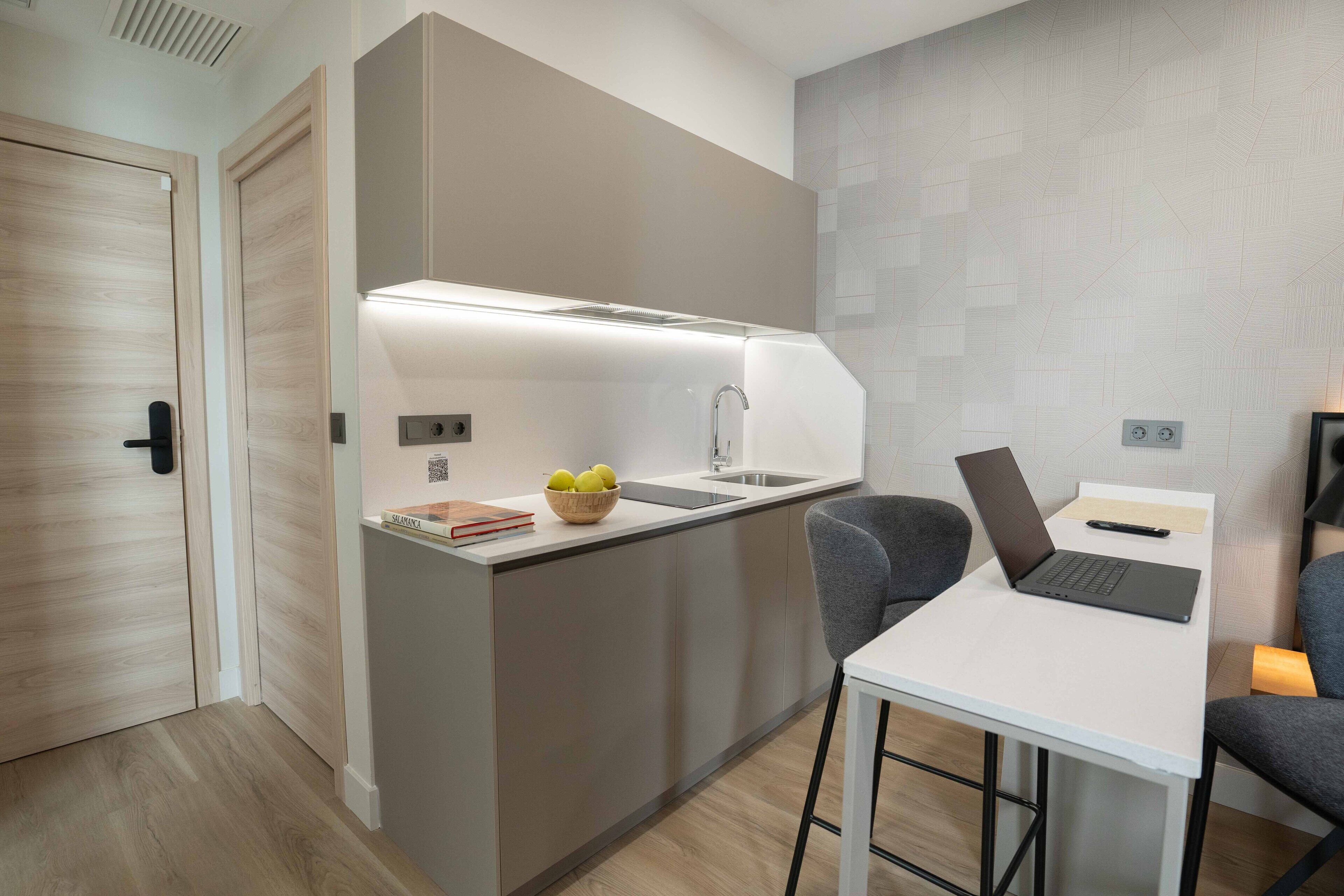 A modern kitchen with a built-in island, a laptop on a small table, and a bowl of fruit on the counter.