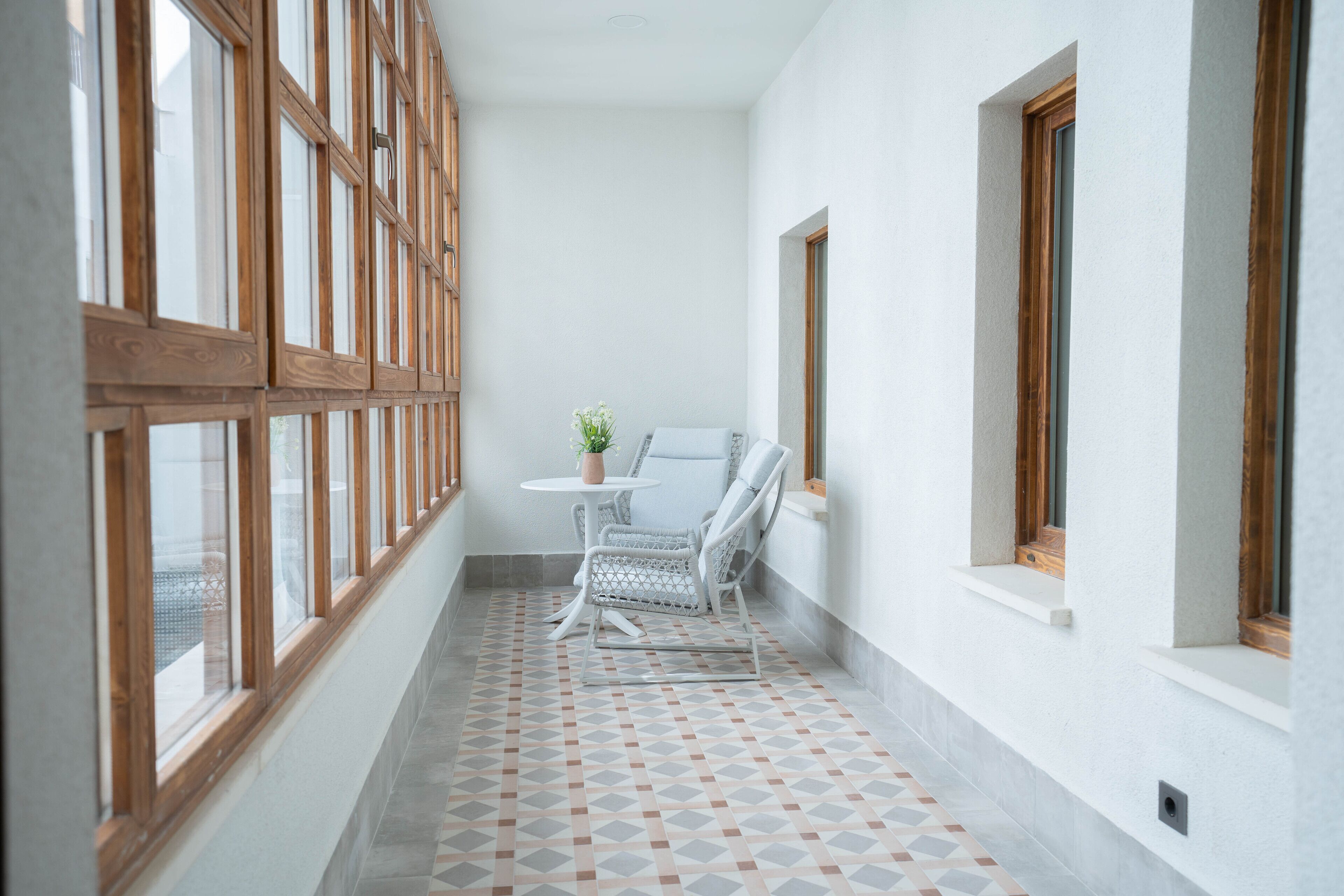 Un pasillo estrecho con suelo de baldosas, una silla blanca, una mesita con planta en maceta y ventanas amplias con marcos de madera.
