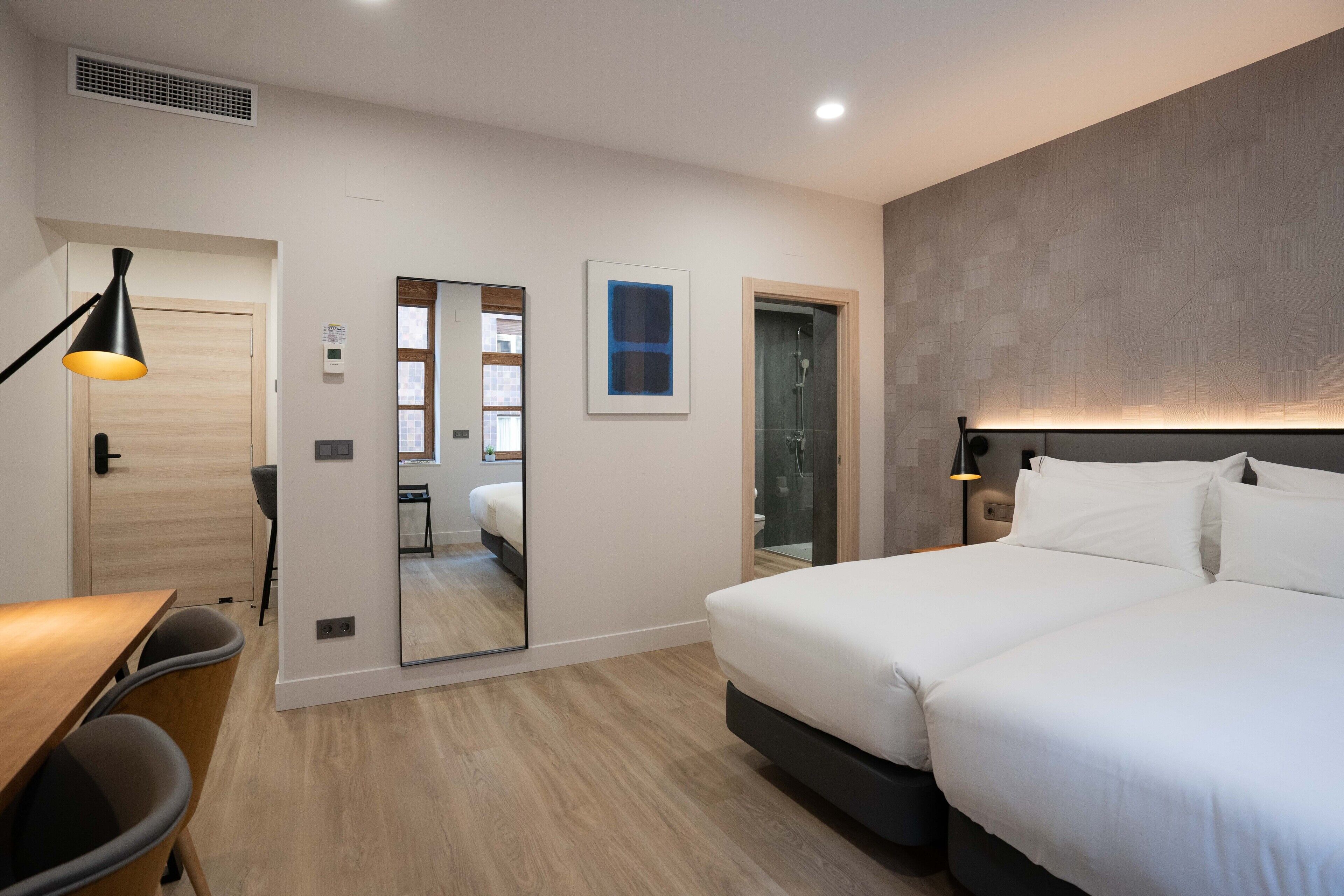 Moderni hotellihuone, jossa on suuri sänky, työpöytä tuolilla, peili ja suihkutila.