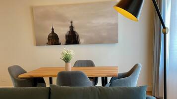 Uma sala de estar moderna com um sofá verde escuro, uma mesa de jantar de madeira com seis cadeiras, um abajur de chão preto e um quadro com uma vista urbana na parede.