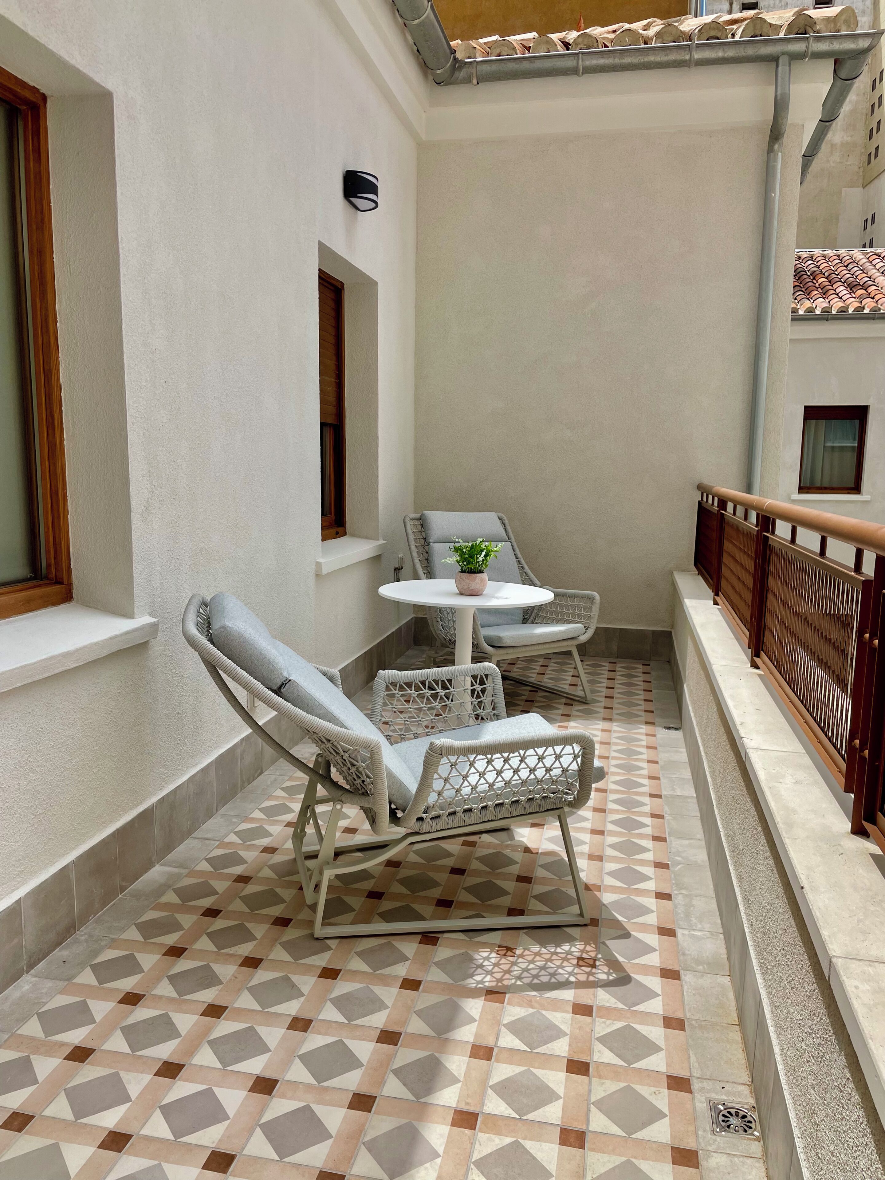 En lille balkon med flisegulv, et hvidt bord med en plante og fletstole.