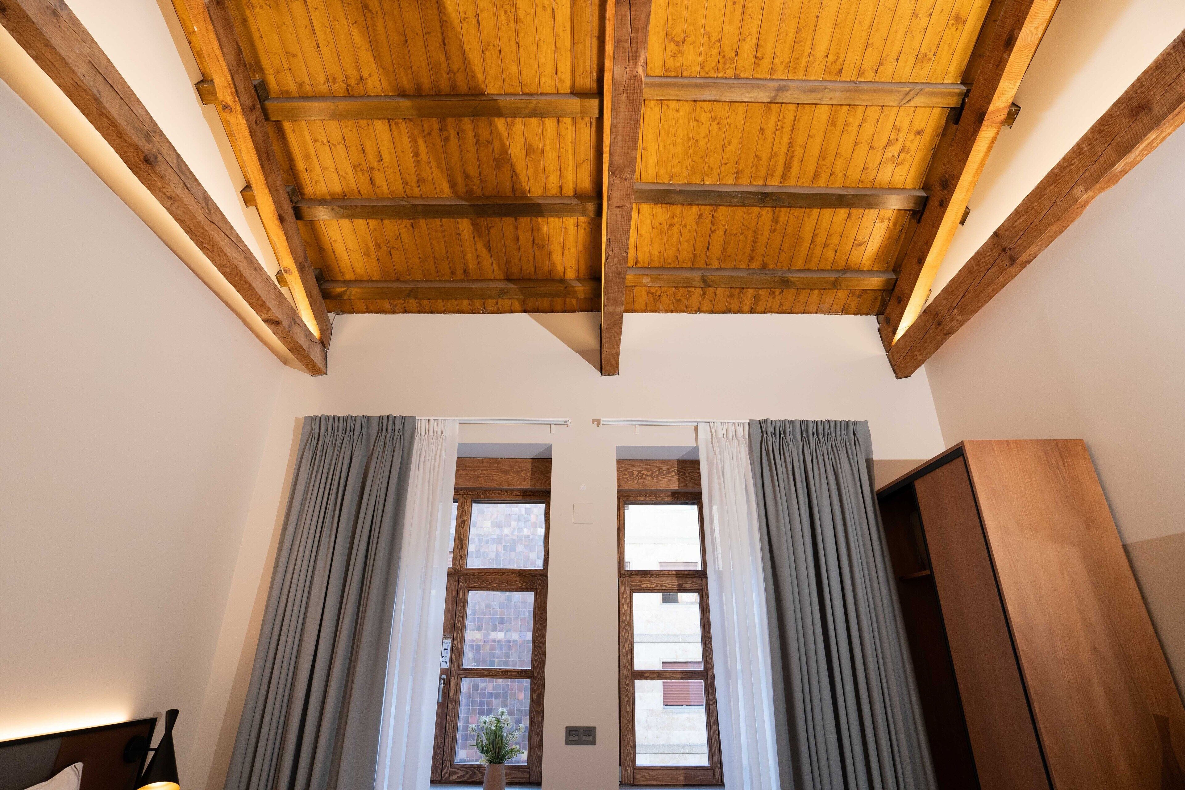 A room with a wooden ceiling, large windows with curtains, and a wooden wardrobe.