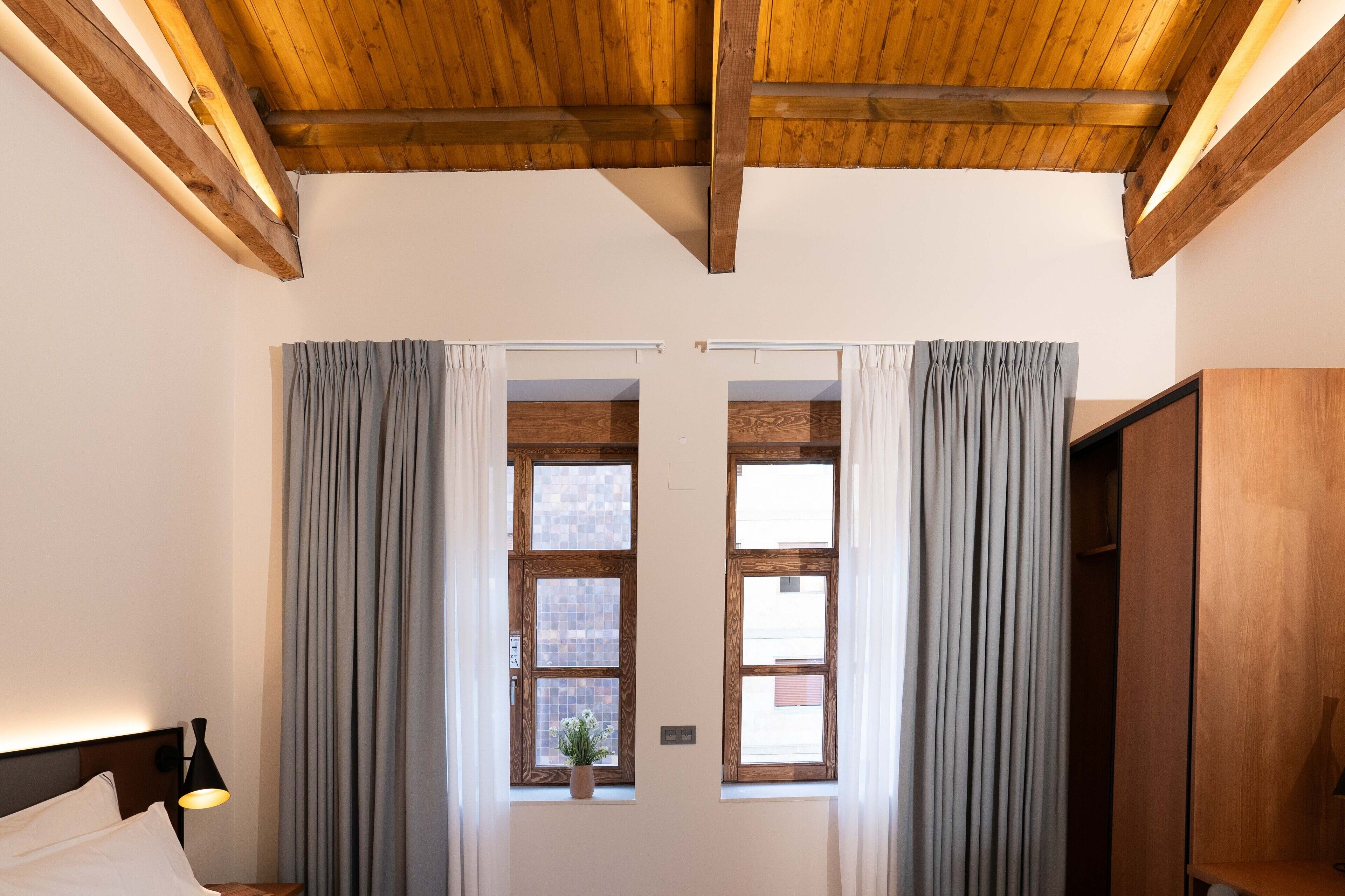 A bedroom with wooden beams, a bed, a nightstand, a lamp, and two windows with curtains.