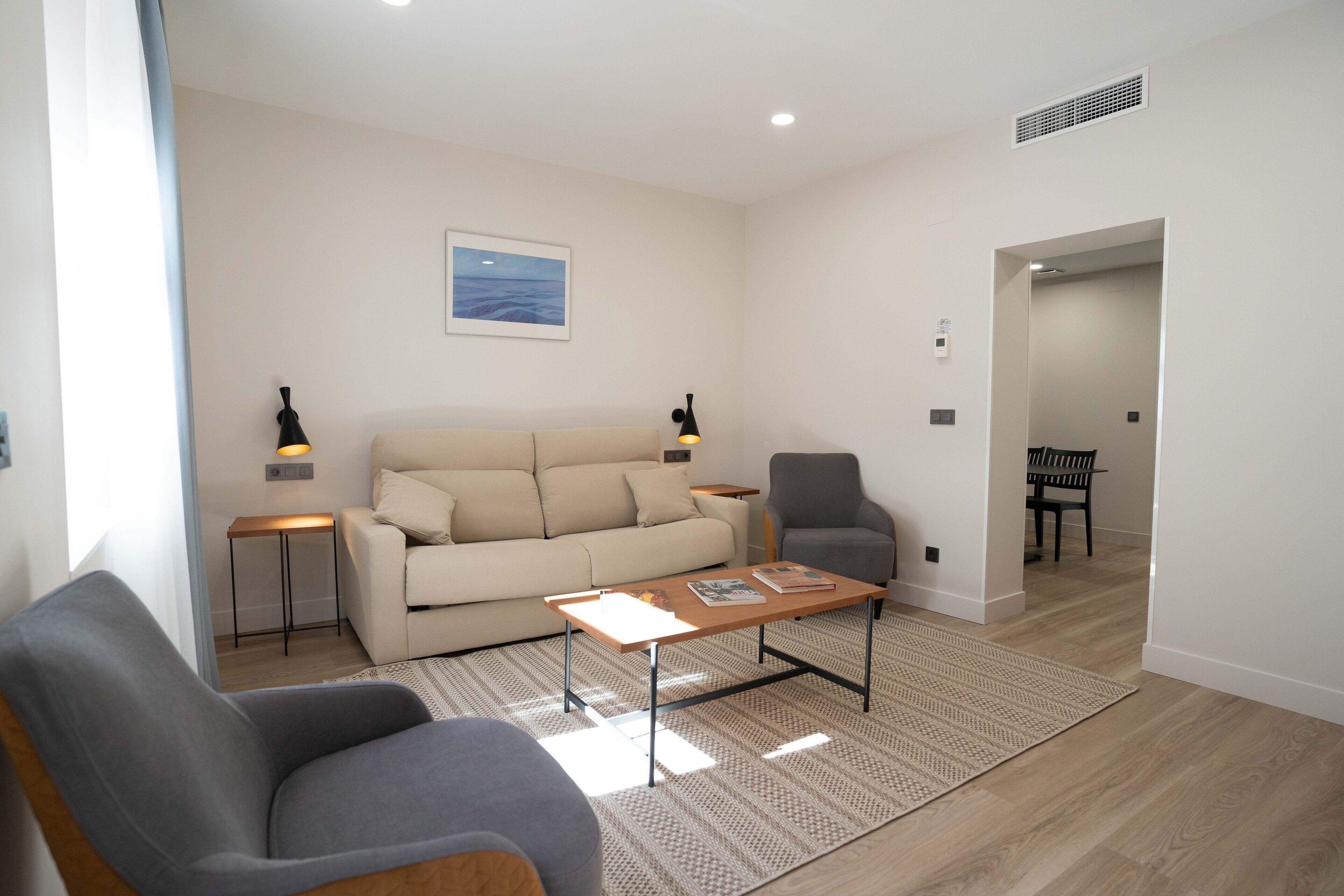 A modern living room with a sofa, two armchairs, a coffee table, and a rug. There is a painting on the wall and a door leading to another room.