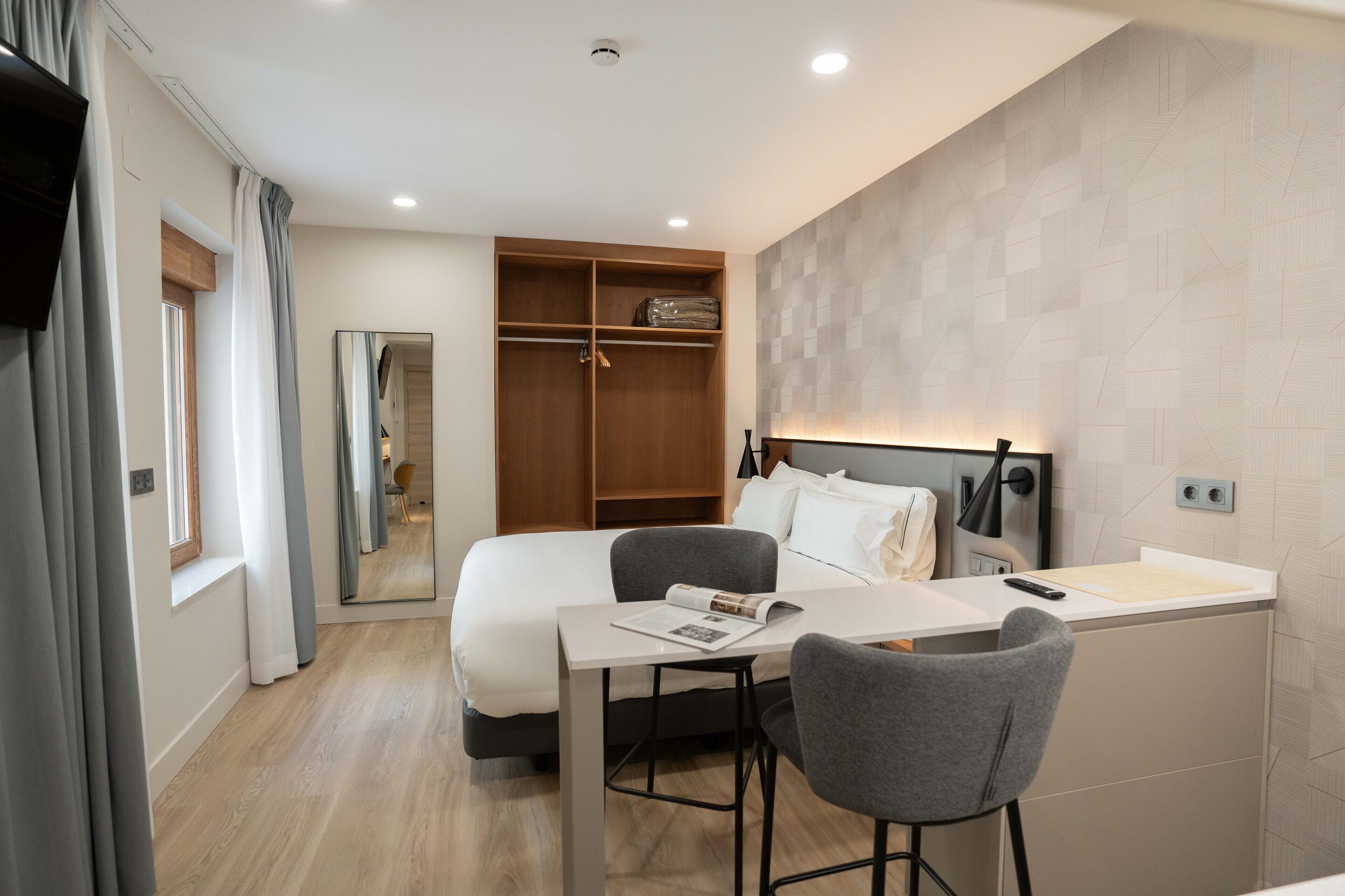 A modern hotel room with a bed, a desk with a chair, a wardrobe, and a mirror.