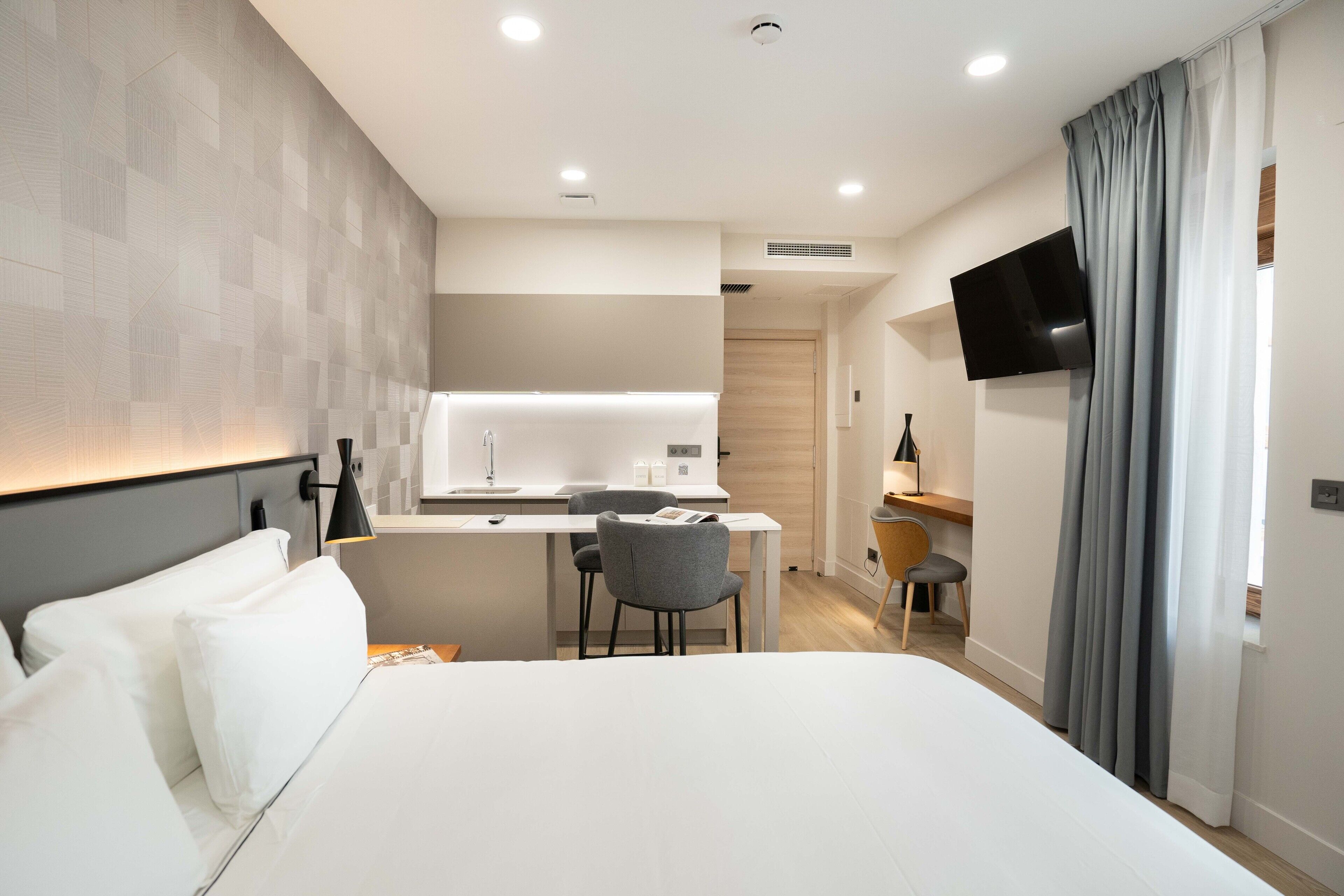 A modern hotel room with a large bed, a desk with a chair, a flat-screen TV mounted on the wall, and a kitchenette area.