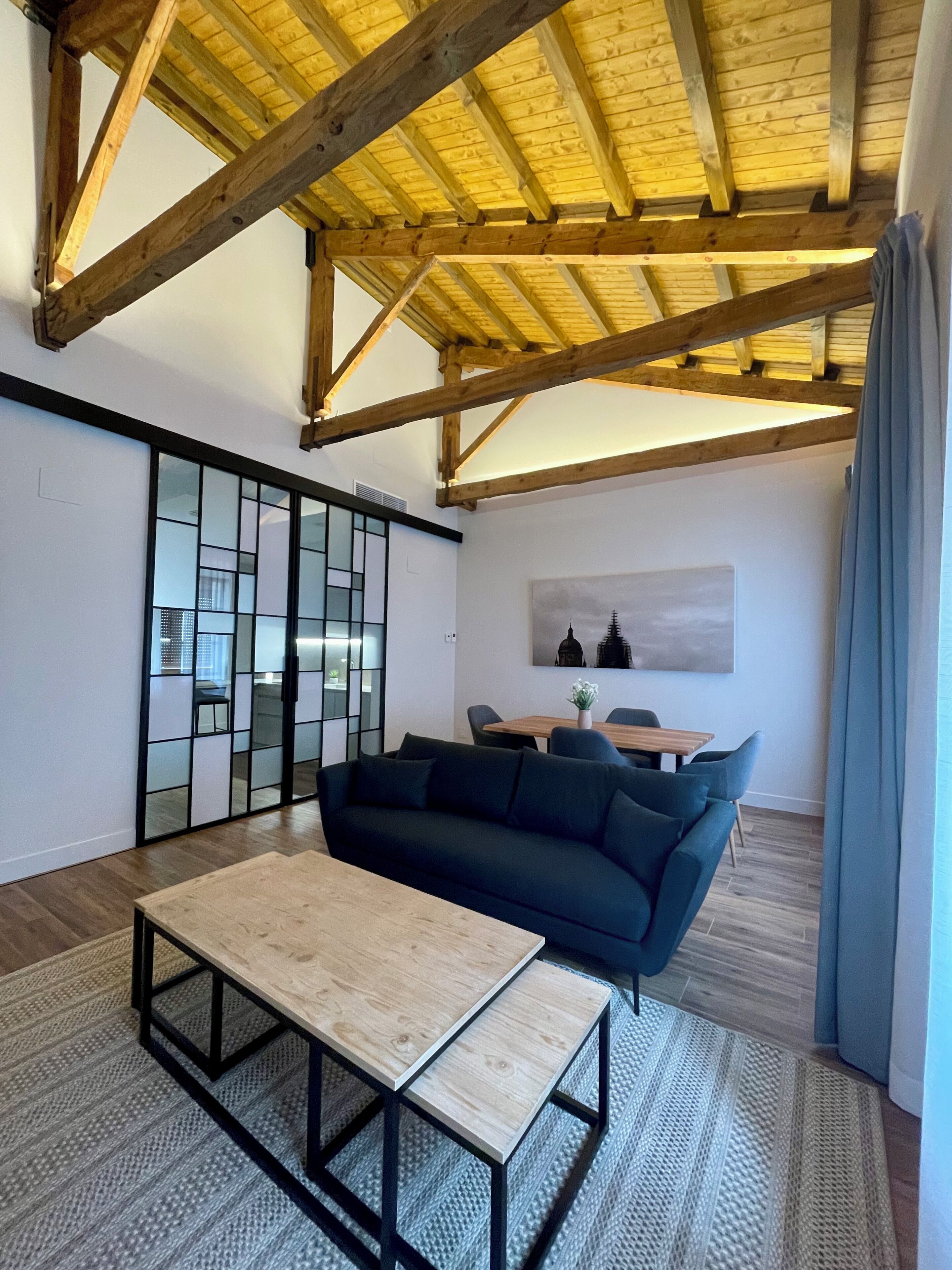A modern living room with a high ceiling, wooden beams, a dark sofa, a wooden coffee table, and a sliding glass door leading to a kitchen.