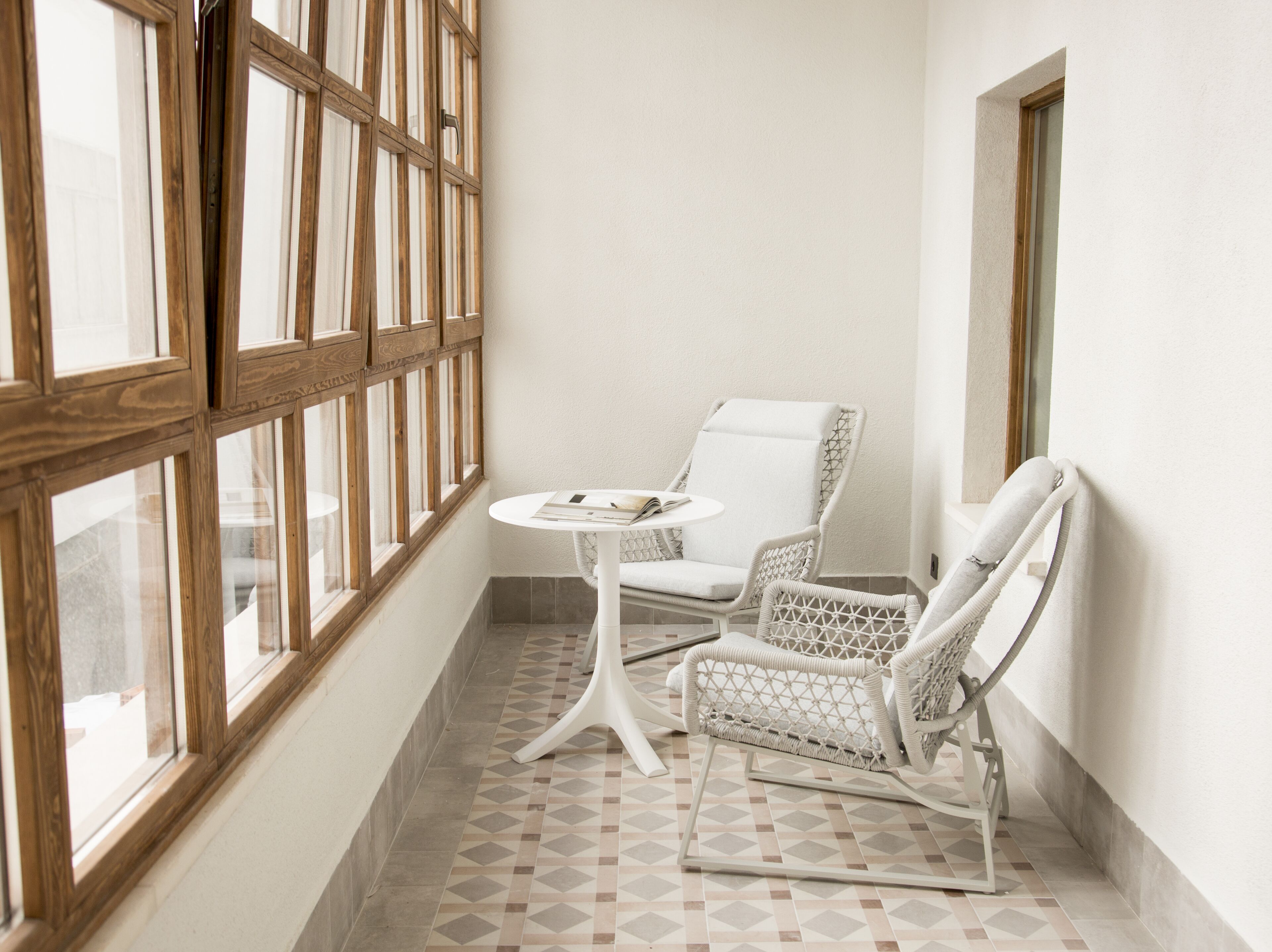 Una habitación de esquina con una silla blanca, una pequeña mesa redonda y un ventanal con marcos de madera.