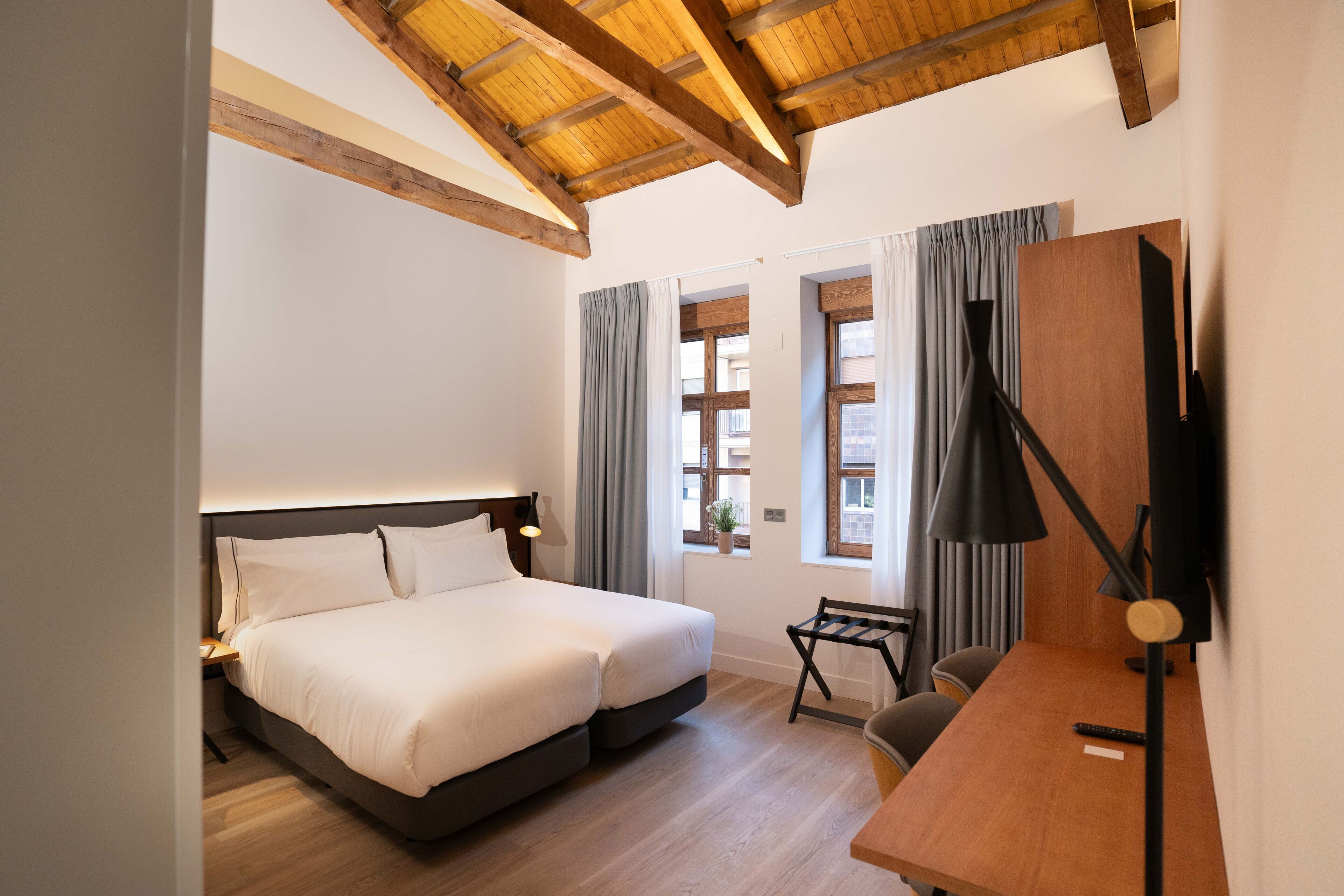 Una camera da letto con un letto, una scrivania e una sedia. Sono presenti finestre con tende, un soffitto con travi a vista e un piccolo tavolo con una pianta.
