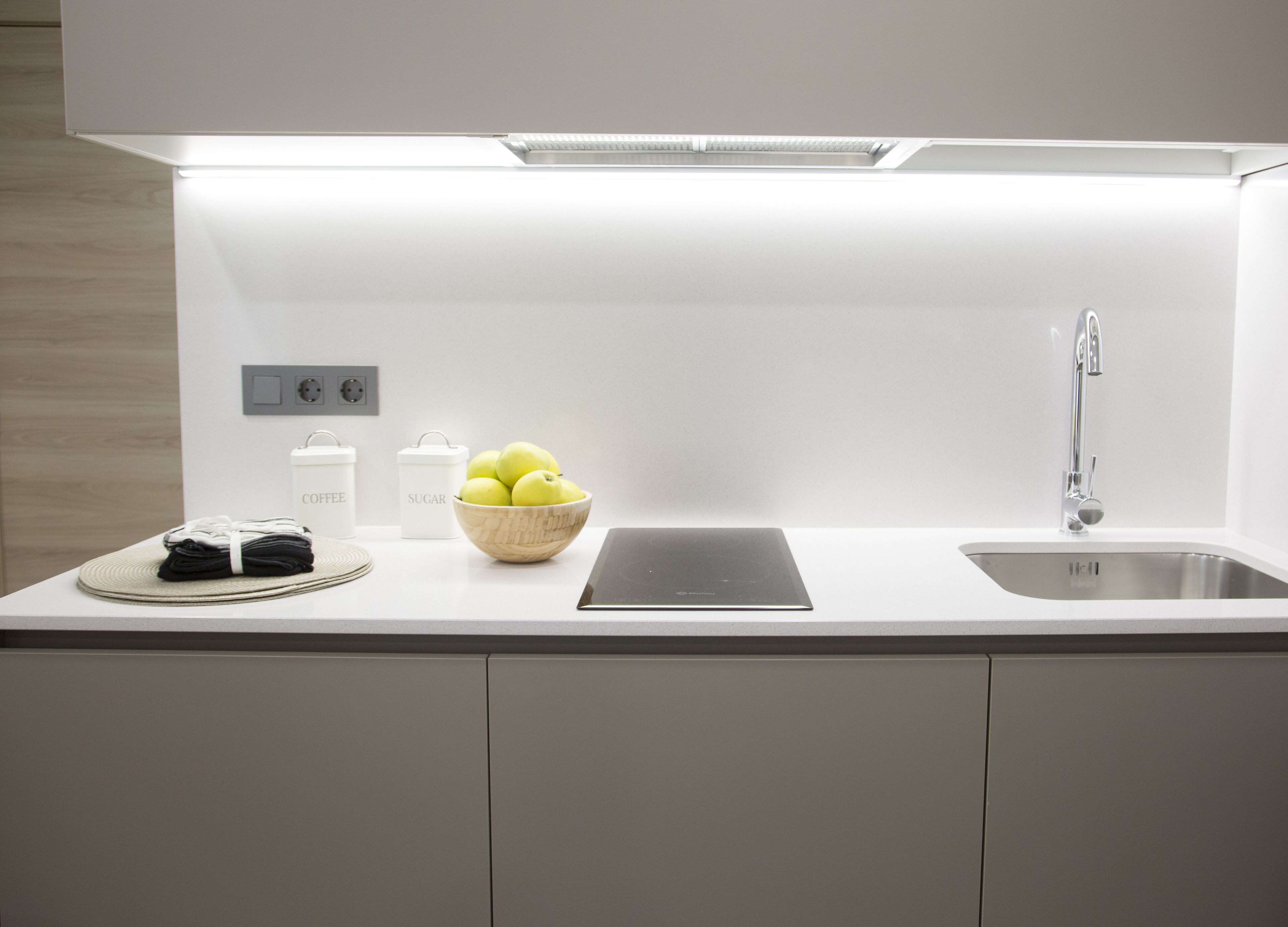 A modern kitchen with a white countertop, a bowl of apples, a coffee maker, and a sink.