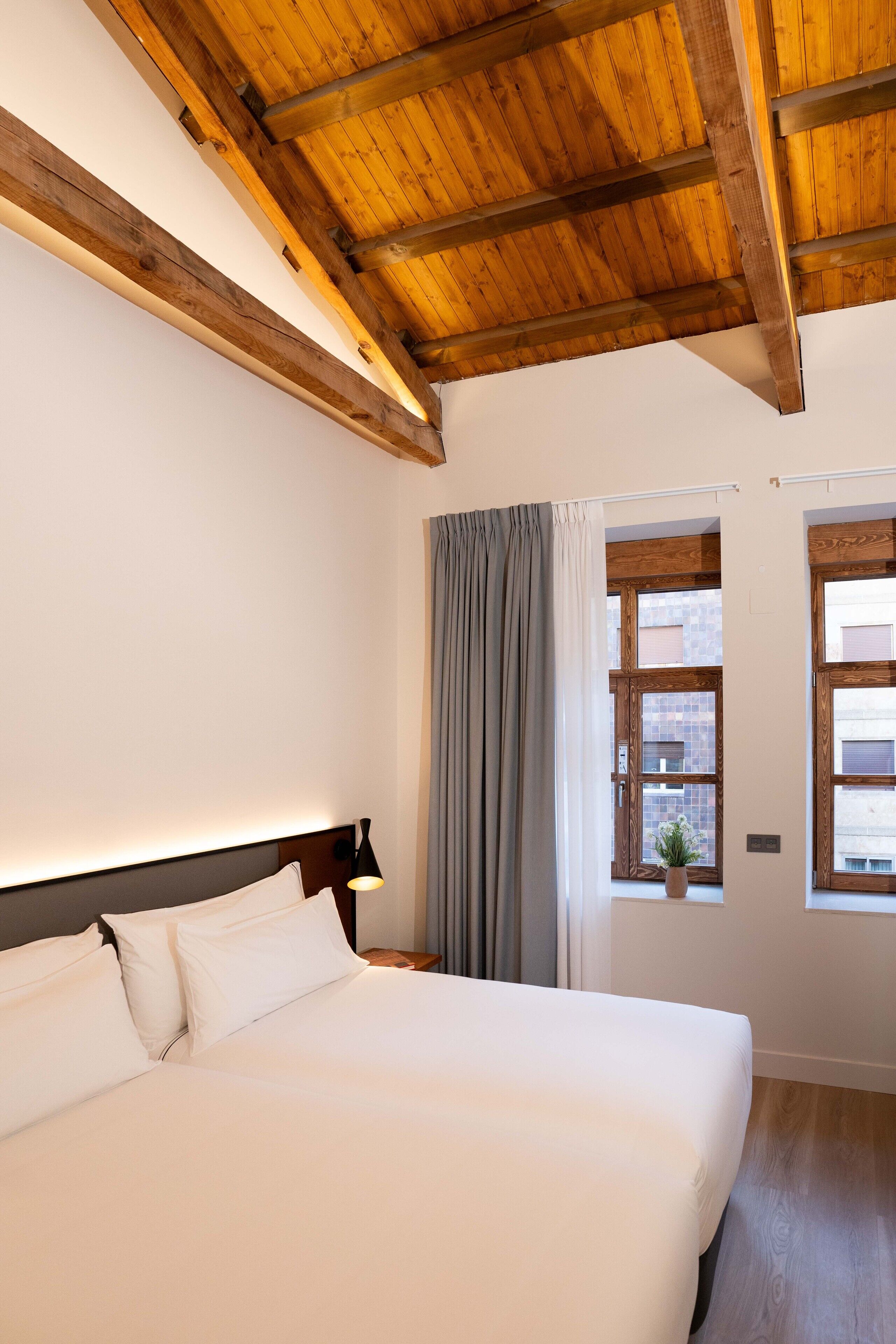 Un dormitorio con techo de madera, una cama grande, una mesita de noche, una lámpara y una ventana con cortinas.
