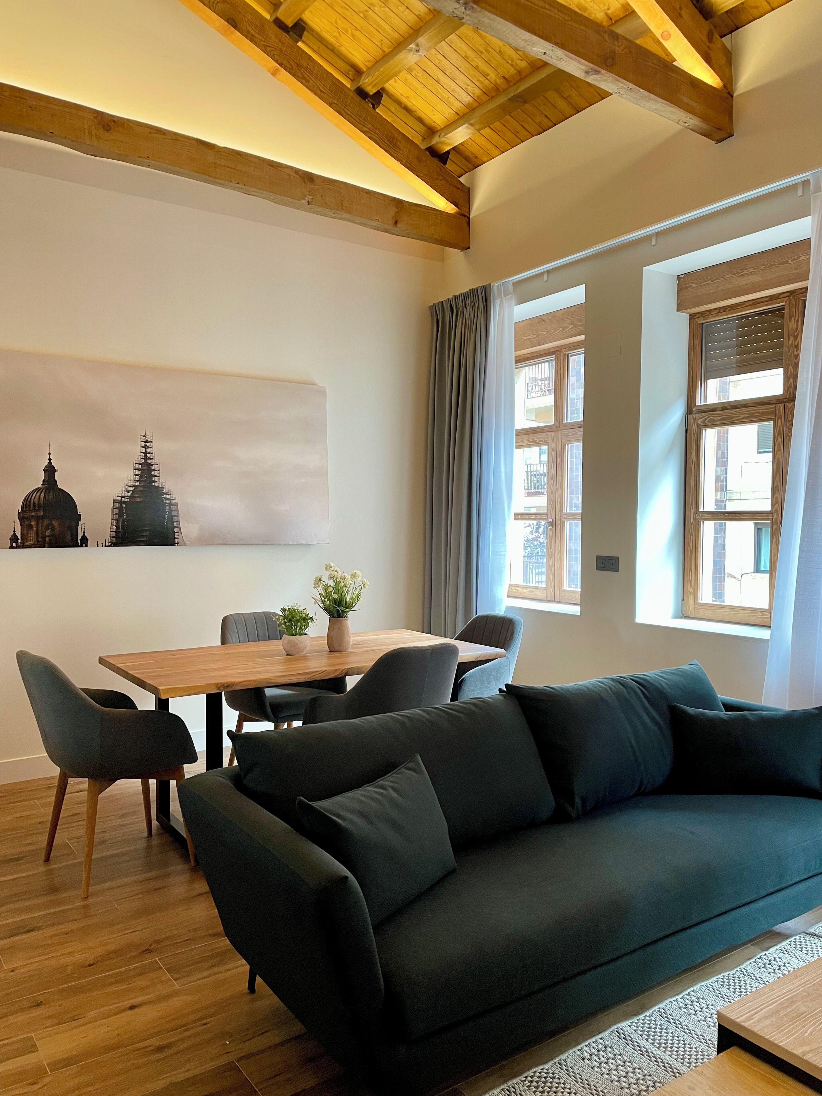 A modern living room with a dark green sofa, wooden dining table, and chairs. A large window with curtains allows natural light.