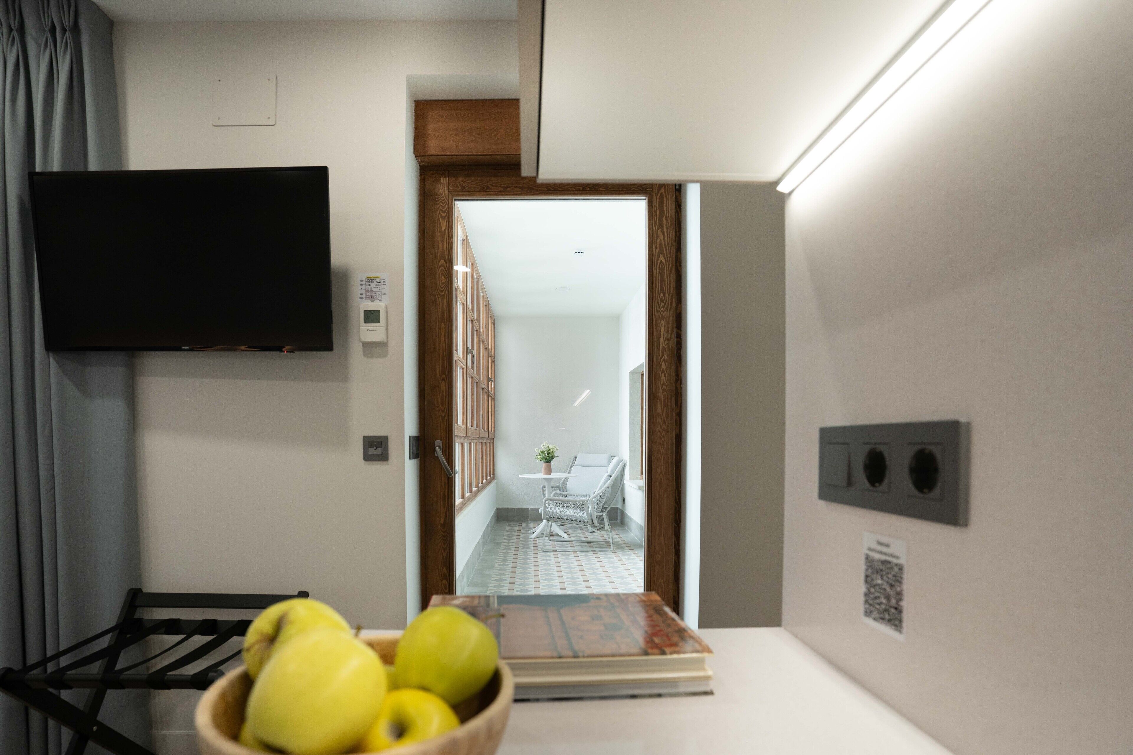 Une chambre d’hôtel dotée d’un téléviseur à écran plat fixé au mur, d’une porte en bois donnant sur un balcon, d’une petite table avec une chaise et d’un saladier rempli de fruits posé sur le comptoir.