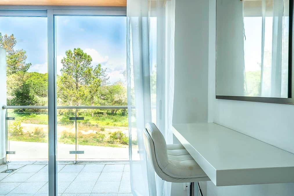Ein modernes Zimmer mit Glastür zu einem Balkon, einem weißen Sessel und einem großen Fenster mit Blick auf Grünflächen und Bäume.