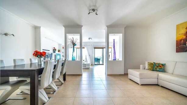 A spacious living area with a dining table and chairs, a white sofa, and a large window providing natural light.