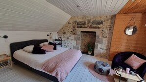 Ein Schlafzimmer mit Kamin aus Stein, einem Bett mit Kopfteil, einem runden Ottomanen und einem kleinen Holztischchen.