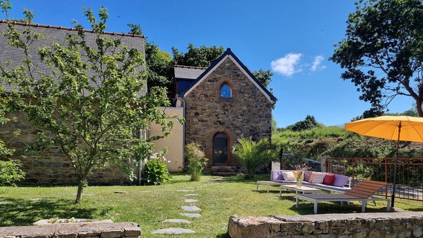 Ein Steinhaus mit einer kleinen Veranda, einem gelben Sonnenschirm und einer Steinmauer mit Stufen zum Eingang.