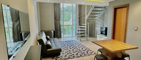 A modern living room with a sofa, a dining table with chairs, a TV mounted on the wall, and a staircase leading to an upper level.