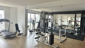 Un gimnasio amplio con diversos equipos de ejercicio, ventanas grandes y un espejo que refleja el espacio.