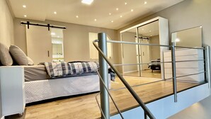Un dormitorio estilo loft moderno con un entrepiso, una cama con almohada a rayas, suelo de madera y una escalera con barandillas metálicas.