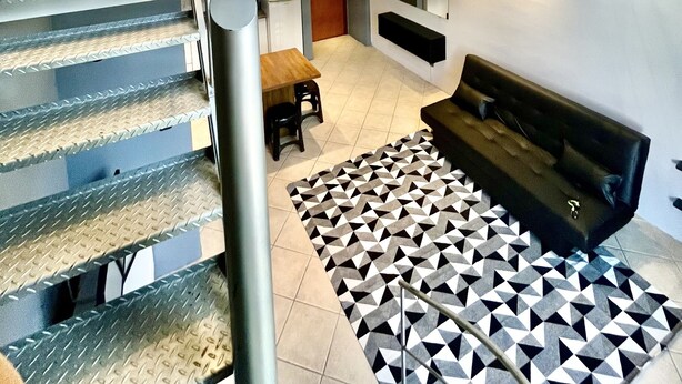 Un interior moderno con una escalera que lleva a una habitación estilo loft, un comedor y una zona de estar con alfombra estampada.