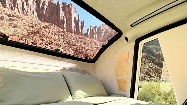 A bed with a quilted cover and a window with a view of rocky cliffs. - Red Rock Base Camps Teardrop Trailer #3. Take your hotel room outside!  (Moab)