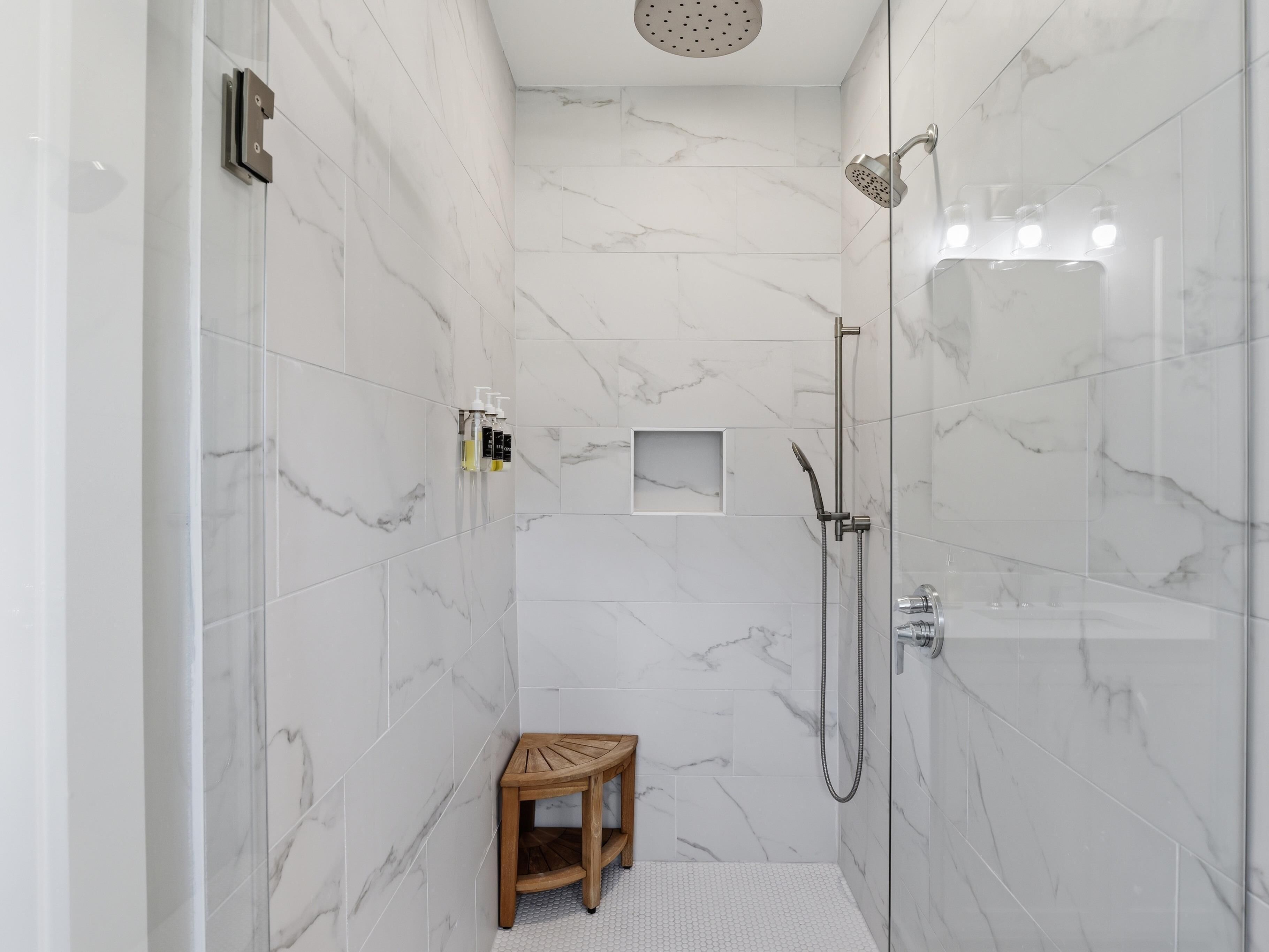 A modern bathroom with white marble tiles, a glass-enclosed shower, a wooden stool, and a small shelf with toiletries.
