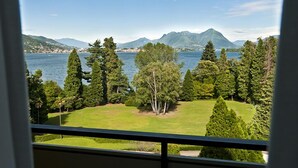 Blick aus einem Fenster auf einen See mit Bergen im Hintergrund, umgeben von Bäumen und einer Rasenfläche.