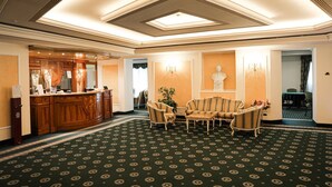 Eine Hotelhalle mit einer Rezeption, einer Sitzecke mit gestreiften Stühlen, einer Kandelaber und einem gemusterten Teppich.