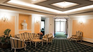 Eine Lobby mit Teppichmuster, gestreiften Sesseln, einer Büstenstatue und einem Kronleuchter.