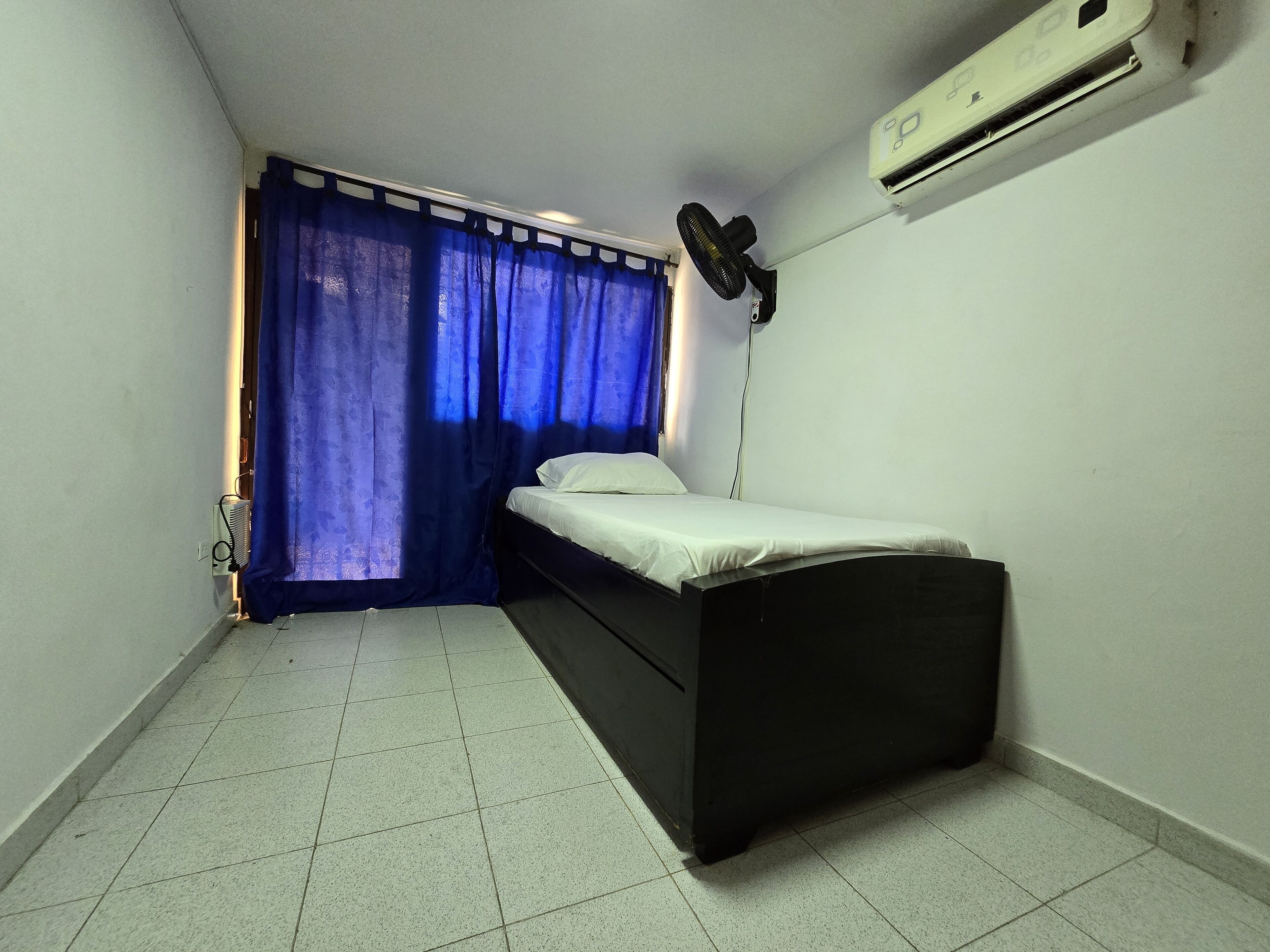 Une petite pièce faiblement éclairée, avec un lit simple, un ventilateur sur pied et un appareil de climatisation fixé au mur.