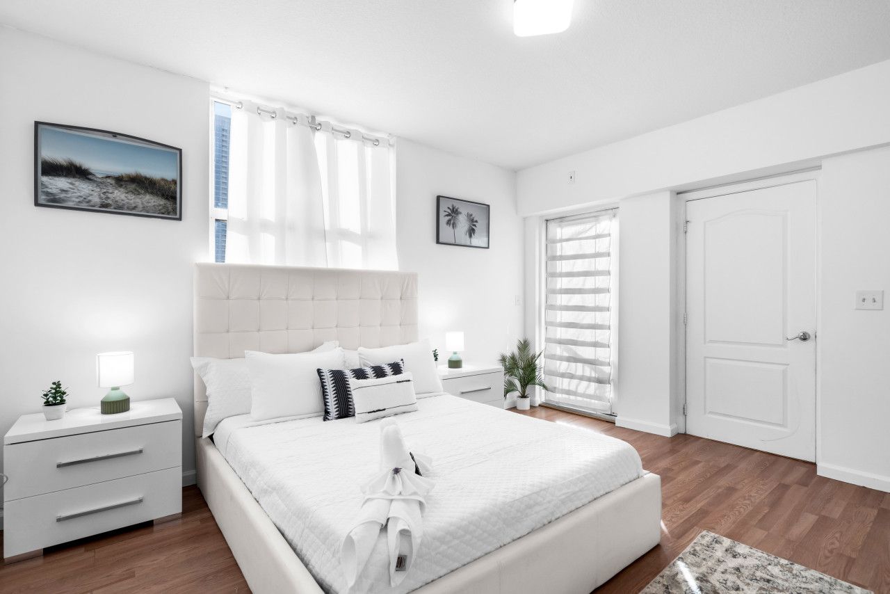 Un dormitorio moderno con una cama grande, cortinas blancas, una mesita de noche con lámpara y un cuadro enmarcado en la pared.