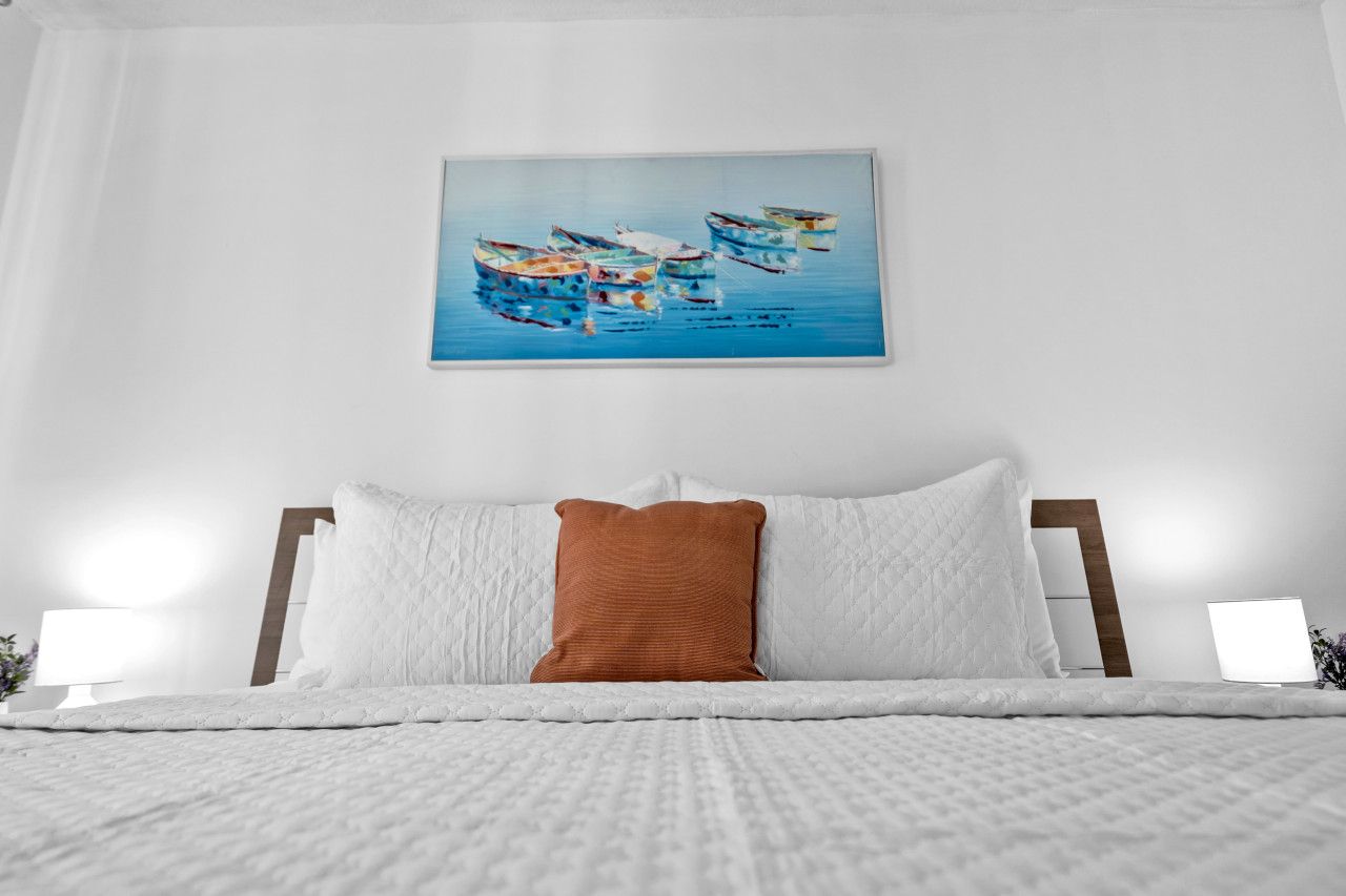 A neatly made bed with a white comforter and a brown pillow, a painting of boats on the wall, and a bedside lamp.