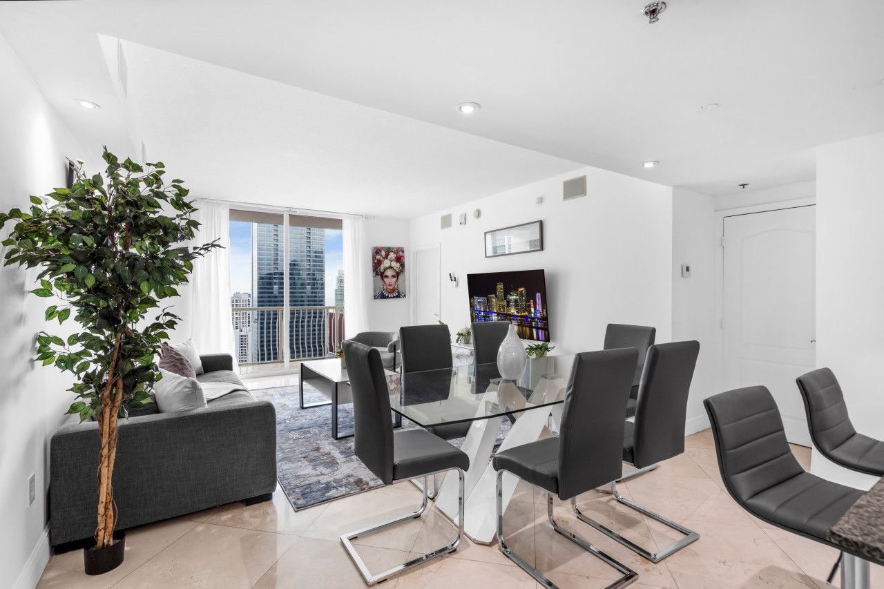 A modern living room with a glass coffee table, black chairs, a grey sofa, a large potted plant, and a city view through the window.