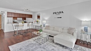 A modern living room with a white sofa, glass coffee table, and a kitchen area with wooden cabinets and white appliances. - Amazing 1 Bed in Brickell key W/ FREE PKG (Miami)