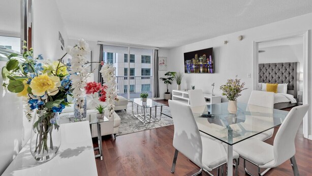 A modern living room with a glass dining table, white sofas, and a large window with a city view. - Amazing 1 Bed in Brickell key W/ FREE PKG (Miami)