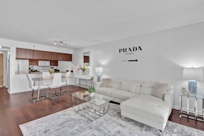 A modern living room with a white sofa, glass coffee table, and a kitchen area with wooden cabinets and white appliances. - Amazing 1 Bed in Brickell key W/ FREE PKG (Miami)